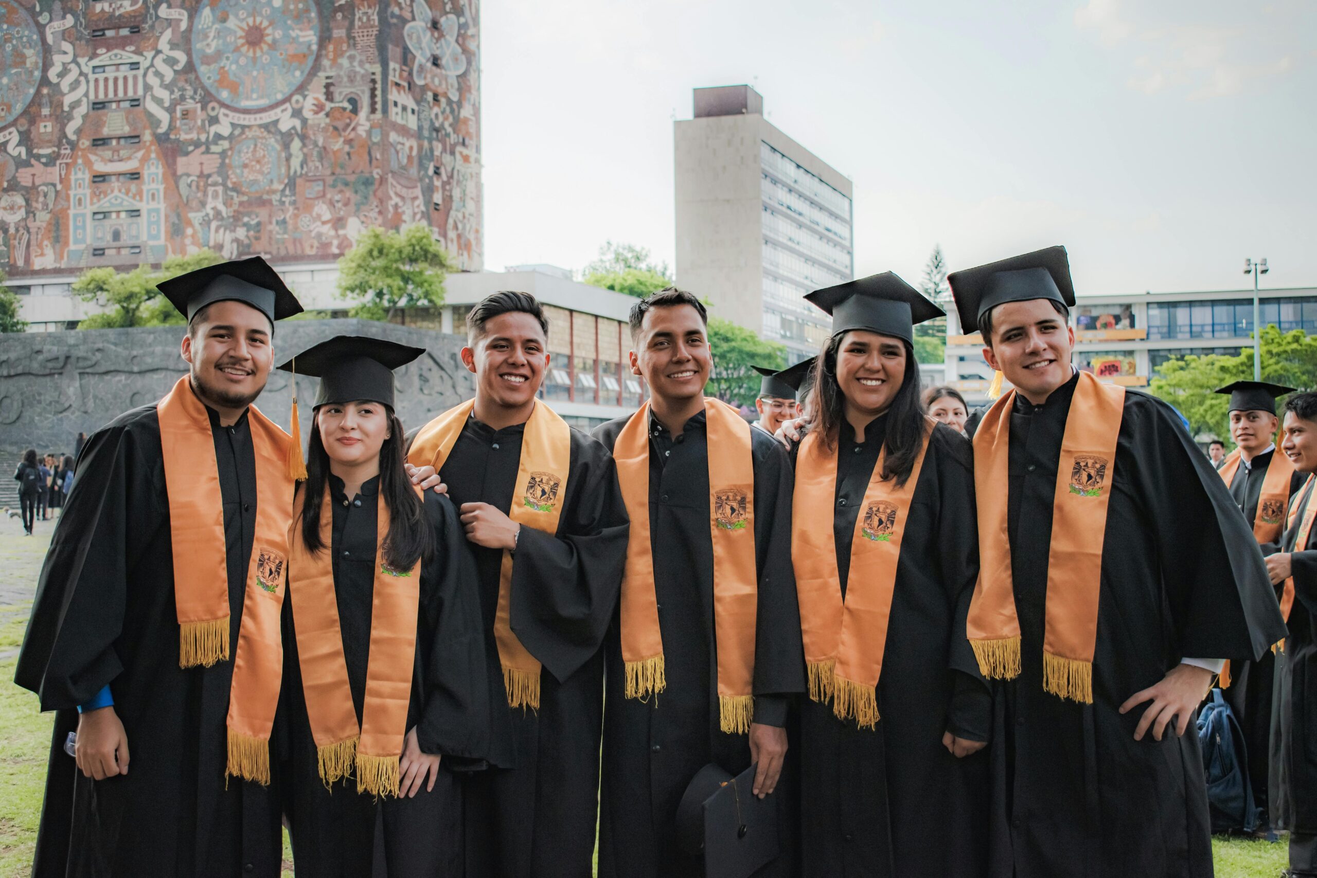 Smiling UNAM graduates in gowns standing together outdoors in front of a campus building.