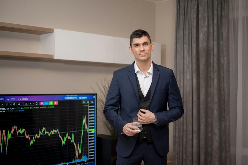 A businessman in a suit stands indoors with a stock market graph on a screen.