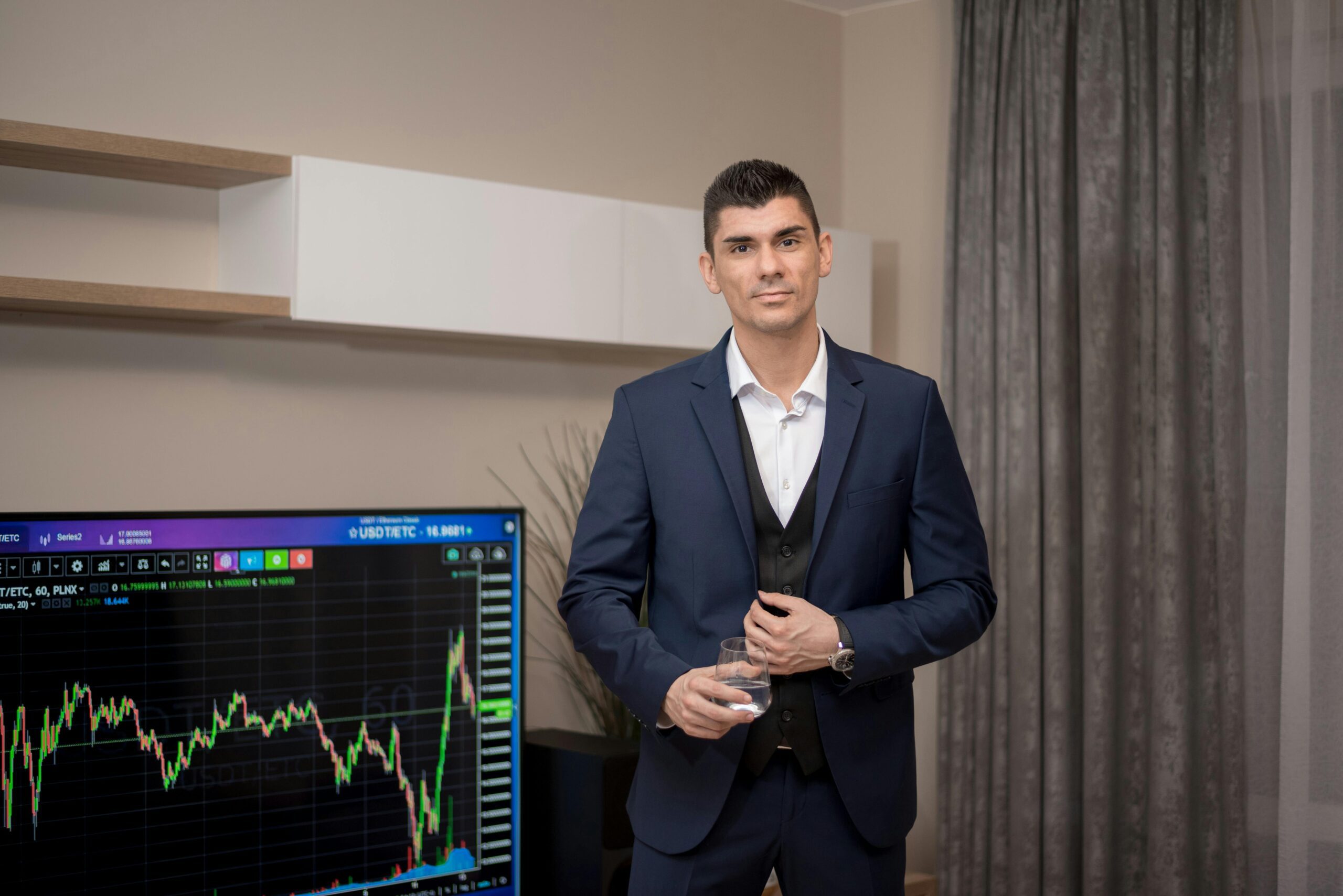 A businessman in a suit stands indoors with a stock market graph on a screen.